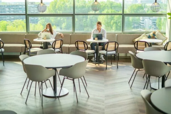 Twee mensen werken op laptops en drinken koffie in een moderne lounge aan de Joop Geesinkweg met uitzicht op groen.